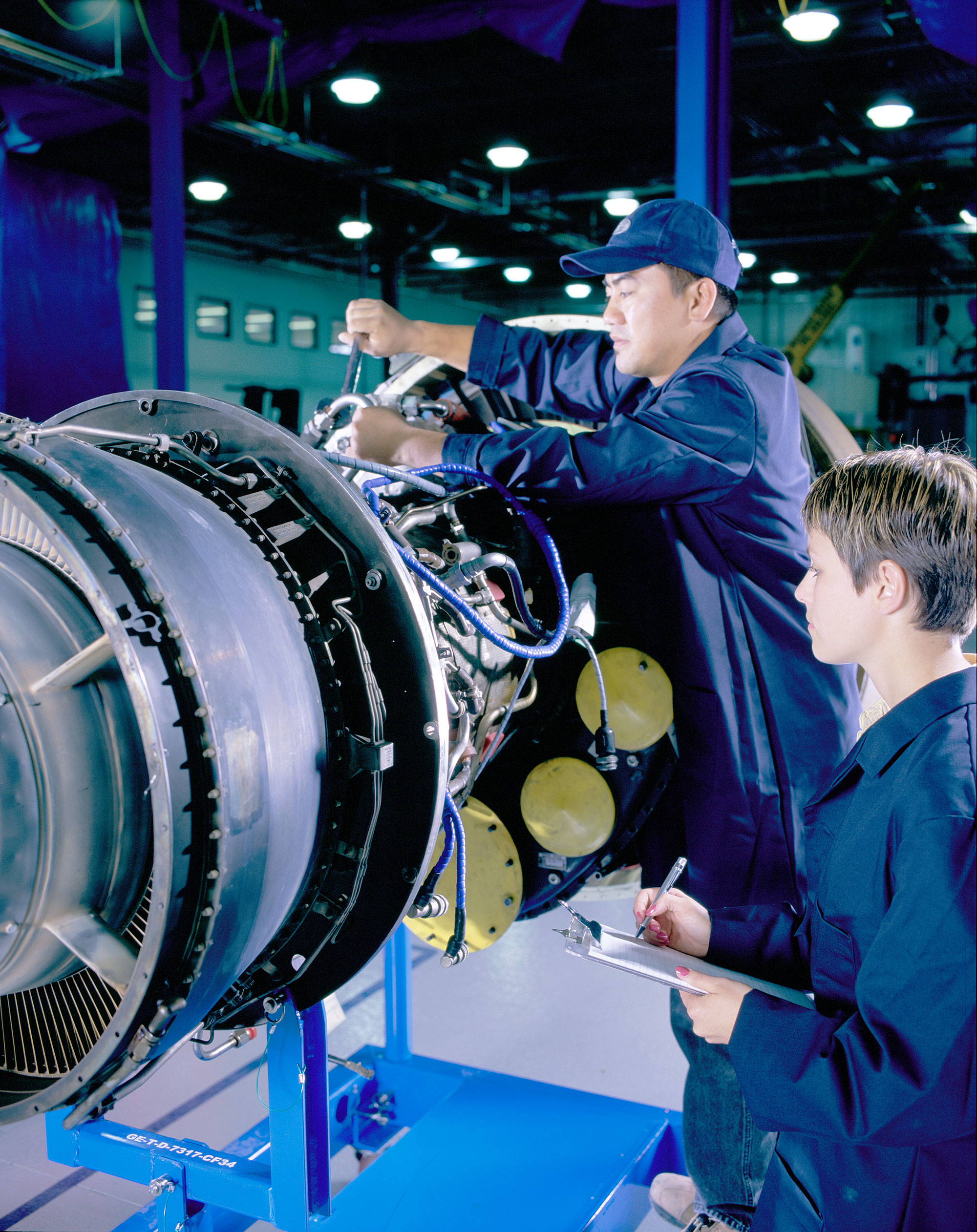 Male worker working on an equipment while a female intern looks on and takes notes.