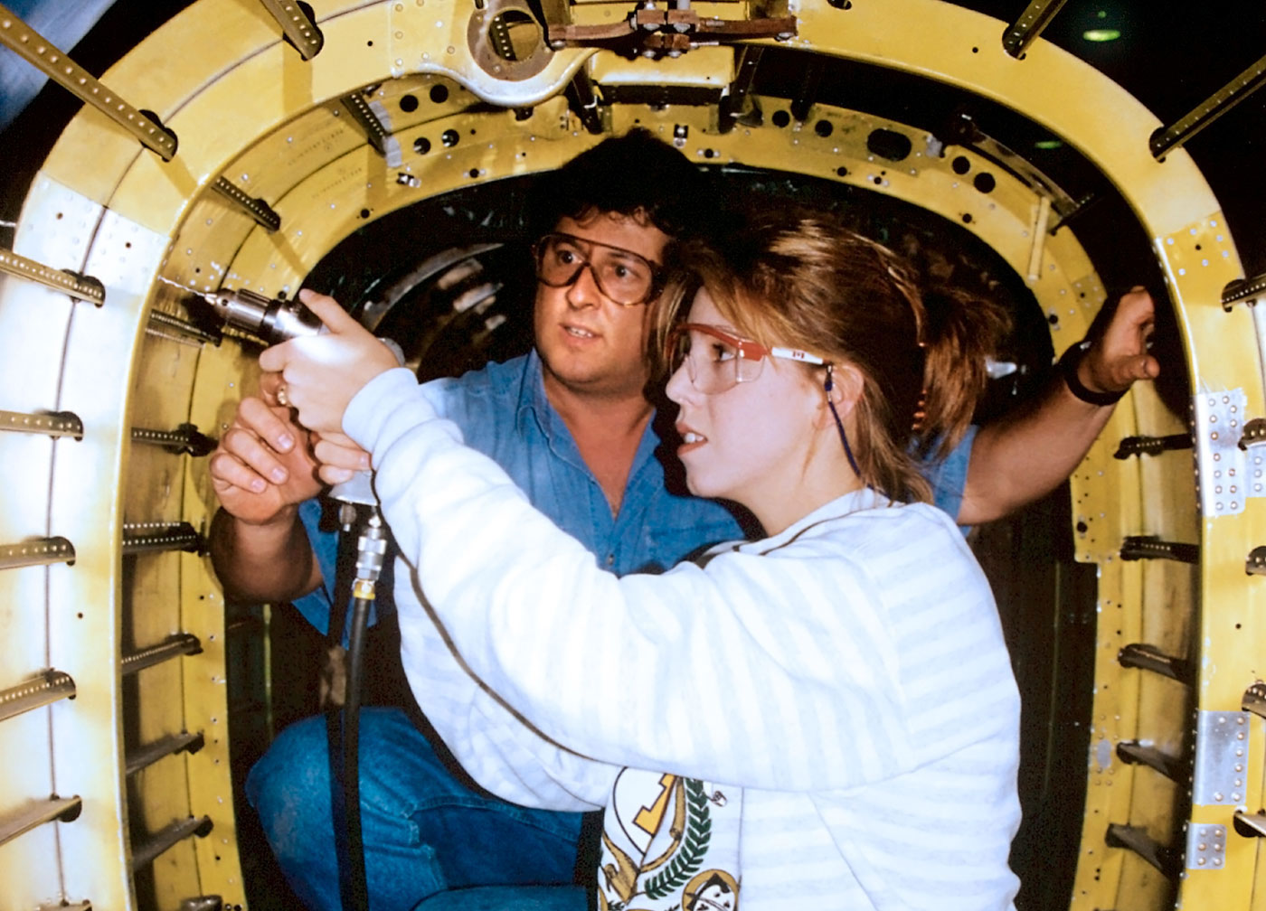 A young female intern operating a tool while a supervisor guides her.